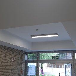 AJ trockenbau - Biały sufit z wnęką i oświetleniem LED w budynku z ceglanymi ścianami. Widoczne drzwi wyjściowe i znaki bezpieczeństwa.