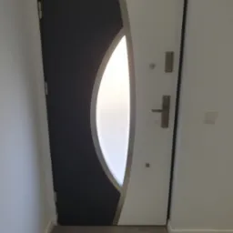 Nowoczesne drzwi wejściowe w kolorze grafitowym i białym z owalnym przeszkleniem, widoczne zamki i klamka, na tle jasnej ściany i podłogi z płytek imitujących kamień.