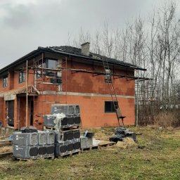 Kacper Torbus - Niedokończony, dwukondygnacyjny dom z czerwonej cegły z częściowo położonym dachem, otoczony rusztowaniami i paletami z materiałami budowlanymi na tle nagich drzew.