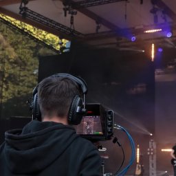 Blue tn sp. j - Operator kamery z tyłu, w słuchawkach, filmuje koncert na scenie z oświetleniem. Widoczny ekran kamery z nagrywanym obrazem muzyka z gitarą.