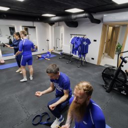 Mocnia - Trening EMS Trener Personalny Tarnów - Trening personalny w studiu: grupa osób w sportowych strojach ćwiczy pod okiem trenera. Widoczne sprzęty: bieżnia, kettlebells, liny TRX. W tle lustra i wieszak z ubraniami.