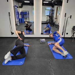 Mocnia - Trening EMS Trener Personalny Tarnów - Dwie kobiety ćwiczą na matach w sali fitness, odbijając się w lustrze. Jedna w czarnym stroju, druga w niebieskim. Tablet z instrukcjami obok. Sprzęt do ćwiczeń w tle.