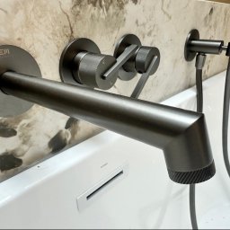 AM design Anna Mrugała - Nowoczesna, ścienna bateria wannowa EMPORIA w matowym antracycie, z oddzielnymi pokrętłami i słuchawką, na tle beżowej ściany imitującej marmur, nad białą wanną Corsan.