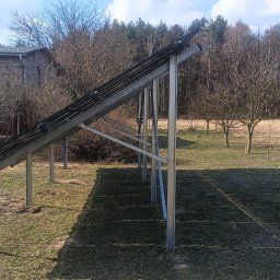 WattProPoland - Montaż paneli słonecznych na metalowej konstrukcji w ogrodzie, widok z boku. W tle budynek i drzewa bez liści. Wiosenna sceneria, błękitne niebo.