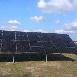 WattProPoland - Czarny panel fotowoltaiczny na metalowej konstrukcji, ustawiony na polu pod błękitnym niebem z chmurami. W tle widoczny dom i drzewa.
