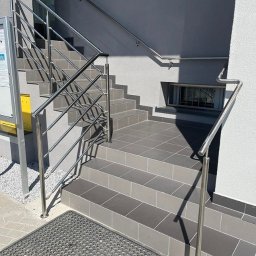 HM Spaw Maciej Stefański - Zewnętrzne schody z szarych płytek prowadzą do budynku. Po obu stronach zamontowano proste, metalowe poręcze ze stali nierdzewnej. Widoczna wycieraczka przed schodami.