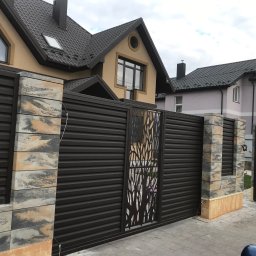 V-I BrukGarden - Nowoczesna brama wjazdowa z paneli w kolorze ciemnego brązu, z dekoracyjnym ażurowym wzorem drzewa, osadzona na słupkach z imitacji kamienia piaskowca.