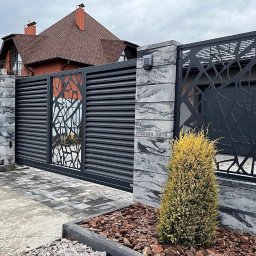 V-I BrukGarden - Nowoczesna brama wjazdowa z poziomych paneli, zdobiona ażurowym wzorem drzewa, osadzona na słupkach z szarego betonu. Ułożona kostka brukowa i ozdobne krzewy.