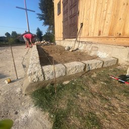 V-I BrukGarden - Budowa obrzeża z kamiennych bloków przy drewnianym budynku. Widoczna szpula sznurka, łopata i pracownik w tle. Prace brukarskie w toku, zarys obrzeża.