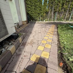 Szymon Podbielski - Umyta kostka brukowa w odcieniach brązu i żółci, prowadząca wzdłuż budynku z klimatyzatorem i zieleni. Widoczne panele słoneczne na dachu.