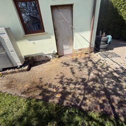 Szymon Podbielski - Częściowo umyta kostka brukowa przed budynkiem z klimatyzatorem i stertą opon. Widoczne mokre i suche fragmenty kostki. Cień drzewa na powierzchni.