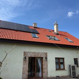 Szymon Podbielski - Dom z czerwonym dachem, oknami dachowymi i panelami słonecznymi. Widoczny komin i klimatyzator na ścianie budynku. Błękitne niebo w tle.