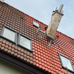 Szymon Podbielski - Czerwona dachówka, okna dachowe i komin z cegły na tle pochmurnego nieba. Widoczne zabrudzenia na dachu i oknach. Ujęcie z dołu, podkreślające strukturę dachu.