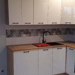 DOMPRO wykończenia wnętrz - Nowoczesna kuchnia z białymi, błyszczącymi szafkami górnymi i dolnymi, drewnianym blatem oraz czarnym zlewem z baterią. Ściana nad blatem wyłożona wzorzystymi płytkami w odcieniach szarości. Jasna...