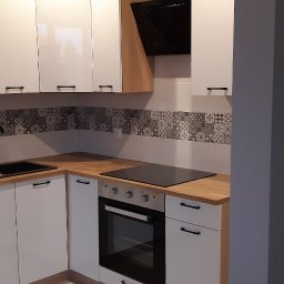 DOMPRO wykończenia wnętrz - Nowoczesna kuchnia w kształcie litery L z białymi, lakierowanymi szafkami, drewnianymi blatami i czarnym sprzętem AGD. Ściana nad blatem wyłożona geometrycznymi płytkami.