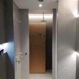 DOMPRO wykończenia wnętrz - Nowoczesna łazienka z szarymi ścianami o fakturze mikrocementu, wbudowanym oświetleniem LED i deszczownicą. Widoczne przyłącza armatury oraz duże kartonowe opakowanie w tle.