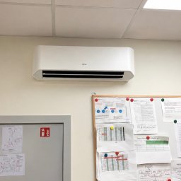 Baltic Klima - Biała jednostka klimatyzatora Fujitsu na jasnej ścianie w biurze, pod sufitem podwieszanym z okrągłym nawiewem i panelem LED. Obok szare drzwi i tablica korkowa z dokumentami.