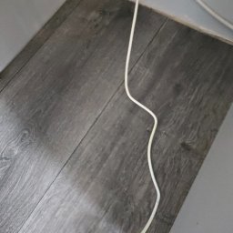 Nova Clean - Szare panele podłogowe z widocznym białym kablem w rogu pomieszczenia, blisko ściany. Widok z góry, oświetlenie naturalne.