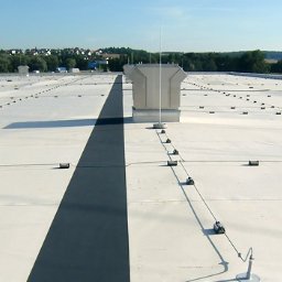 GromOFF - Biały dach budynku z zainstalowaną siatką odgromową i zwodami pionowymi. Widok z góry, w tle zieleń i zabudowa miejska.