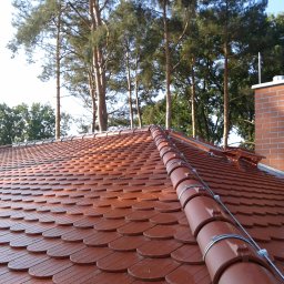 GromOFF - Czerwona dachówka karpiówka z zamontowaną instalacją odgromową, widoczny komin z cegły i drzewa w tle. Metalowe uchwyty mocujące przewód odprowadzający.