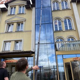 Panel Cleaner - Mycie szklanej fasady budynku z użyciem teleskopowego kija z myjką. Widoczni dwaj mężczyźni obserwujący pracę. Budynek z napisem 'Restaurant'.