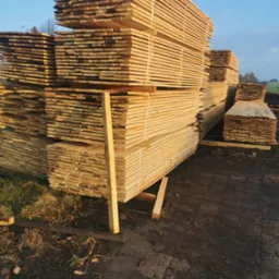 Stosy świeżo przetartego drewna budowlanego ułożone na zewnątrz, zabezpieczone białymi taśmami, z widocznymi elementami podkładu i fragmentami trawy.