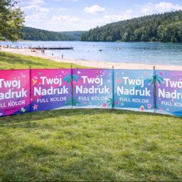 Dream Factory 24 - Parawan plażowy z kolorowym nadrukiem 'Twój Nadruk FULL KOLOR' na tle jeziora i plaży. Widoczne drzewa i osoby w oddali. Reklama na trawie.