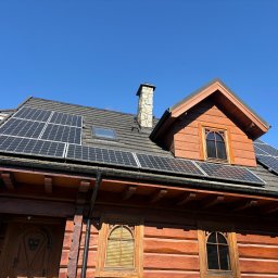 Nowa Energia Maciej Szerechan - Drewniany dom z panelami słonecznymi na dachu, widoczne okna z ozdobnymi ramami i komin z kamienia. Błękitne niebo w tle, ujęcie z dołu.
