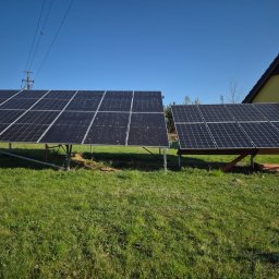 Nowa Energia Maciej Szerechan - Dwa panele fotowoltaiczne na trawie, jeden na metalowych nogach, drugi na drewnianej konstrukcji. W tle dom i słupy energetyczne pod błękitnym niebem.
