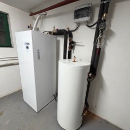 Nowa Energia Maciej Szerechan - Biała pompa ciepła York i zbiornik akumulacyjny w piwnicy, widoczne rury, zawory i skrzynka elektryczna na ścianie. Szare podłoże, surowy wystrój.