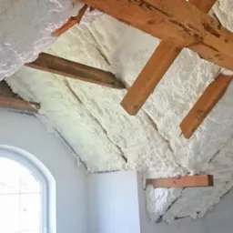 Poddasze z drewnianą konstrukcją i ociepleniem z pianki PUR, widoczne białe okno. Izolacja natryskowa na skośnych ścianach, detale drewnianych belek.