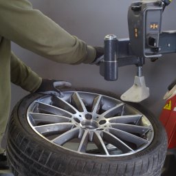 DEWIZA PLUS Monika Bednarska - Opona na montażownicy z felgą aluminiową, obsługiwana przez osobę w rękawicach, w warsztacie. Widoczny fragment maszyny i opona marki Bridgestone.