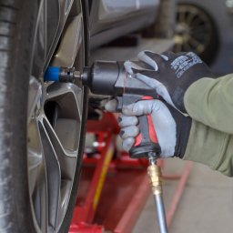 DEWIZA PLUS Monika Bednarska - Koło samochodu na podnośniku, dokręcane kluczem pneumatycznym przez mechanika w rękawicach roboczych. Widoczny przewód pneumatyczny i fragment podwozia.