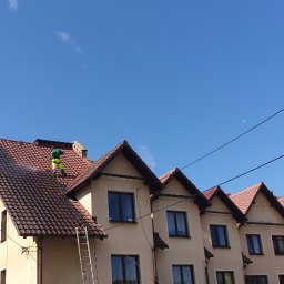 Profi-mal Alicja Wątroba - Pracownik w zielonym kombinezonie czyści dach domu z brązową dachówką za pomocą myjki ciśnieniowej. Na dachu drabina. Błękitne niebo w tle.