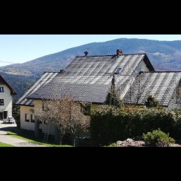 Profi-mal Alicja Wątroba - Dom z ciemną dachówką na tle gór. Na dachu widoczna osoba podczas pracy. Drabina oparta o ścianę. Ujęcie w słoneczny dzień.