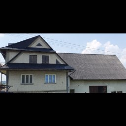 Profi-mal Alicja Wątroba - Dwukondygnacyjny dom z ciemnym dachem i jasną elewacją, widoczny z poziomu gruntu w słoneczny dzień. Dach z blachy trapezowej. Budynek po renowacji.