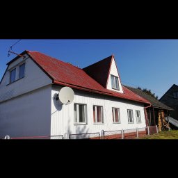 Profi-mal Alicja Wątroba - Biały dom z czerwonym dachem i anteną satelitarną na ścianie bocznej, widziany w słoneczny dzień. Widoczne okna i fragment ogrodzenia.