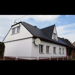 Profi-mal Alicja Wątroba - Dom jednorodzinny z ciemnym dachem i białą elewacją, widoczny z boku. Na ścianie numer 57 i antena satelitarna. Czerwone elementy ogrodzenia.