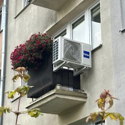 Cooling Service Łukasz Zawiłowicz - Biały klimatyzator Haier zamontowany na ścianie budynku obok balkonu z czerwonymi kwiatami. Widok z dołu, z elementami roślin na pierwszym planie.