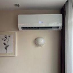 Cooling Service Łukasz Zawiłowicz - Biały klimatyzator Haier na ścianie w pokoju, obok obrazu z motywem kwiatowym i lampy ściennej. Widoczny fragment okna z firanką.