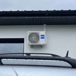 Cooling Service Łukasz Zawiłowicz - Biała jednostka zewnętrzna klimatyzacji Haier zamontowana na elewacji budynku, widoczna zza mokrego dachu samochodu w pochmurny dzień. Minimalistyczny design.