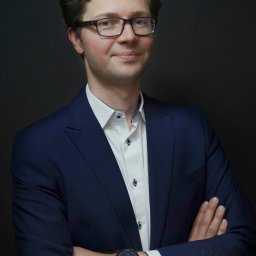 Aleksander Banacki - Portret mężczyzny w okularach i granatowym garniturze na ciemnym tle, z założonymi rękami i widocznym zegarkiem. Uśmiechnięty, profesjonalny wizerunek.
