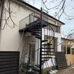 Tomasz Szcześniak - Zewnętrzne, metalowe schody spiralne prowadzące na balkon z czarną balustradą. Widoczne drabiny i narzędzia sugerują trwający montaż przy elewacji domu.