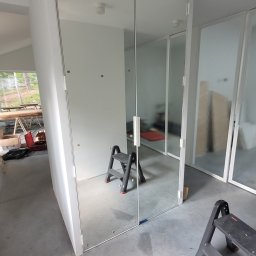 Tomasz Szcześniak - Nowoczesna szklana ścianka działowa z metalowymi ramami, widoczna drabina malarska, surowy beton na podłodze, minimalistyczny design wnętrza.
