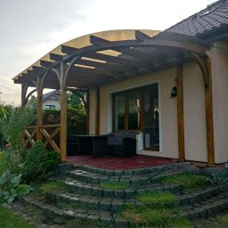 Mateusz Obi - Łukowata pergola drewniana z półprzezroczystym zadaszeniem, połączona z jasnym domem. Na tarasie z czerwonej kostki brukowej ciemne meble ogrodowe, obok kamienne schody i zieleń.
