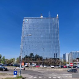 Ankor Biuro Nieruchomości Rybnik - Szklany biurowiec z logo na szczycie, odbijający otoczenie, widoczny z poziomu ulicy z przejeżdżającymi samochodami w słoneczny dzień.