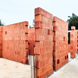 Budowy dla Ciebie - Wznoszone ściany z czerwonej ceramiki poryzowanej na fundamencie. Widoczne zbrojenia pionowe. Budowa w toku, tło stanowią drzewa.