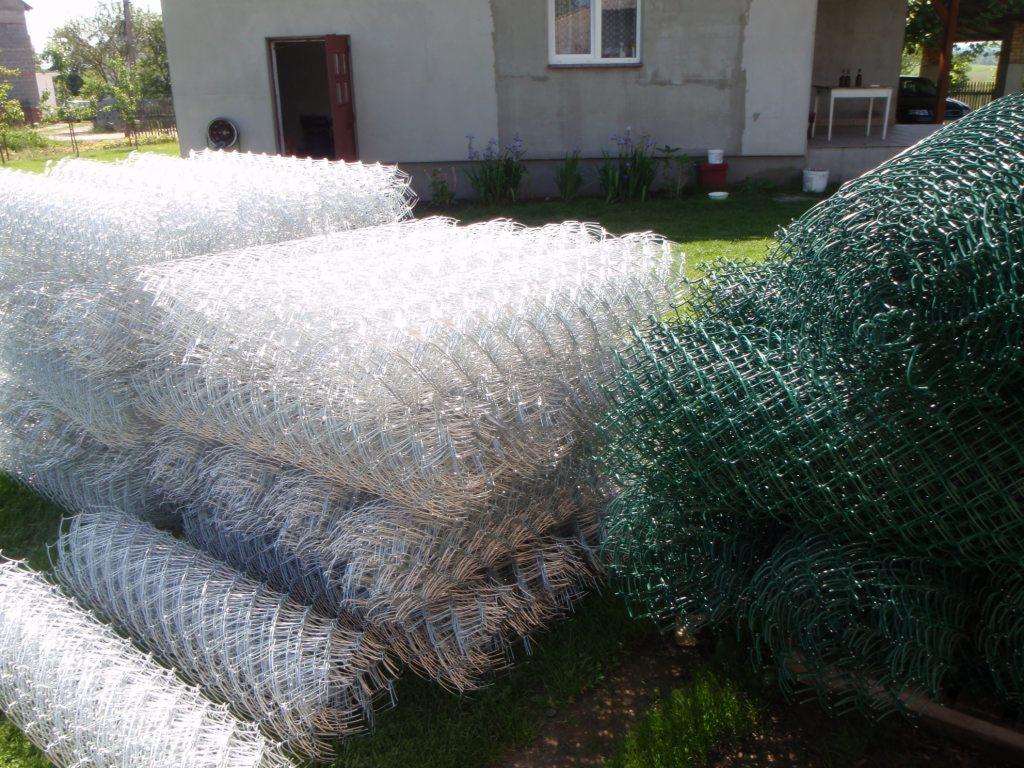 Stosy zwiniętej siatki ogrodzeniowej: ocynkowanej i powlekanej na zielono, leżące na trawie przed budynkiem z otwartymi drzwiami.