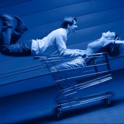 Szelest - Para w niebieskiej tonacji: mężczyzna pcha wózek sklepowy z leżącą kobietą, oboje uśmiechnięci, dynamiczne ujęcie z rozmytym tłem sugerujące ruch.