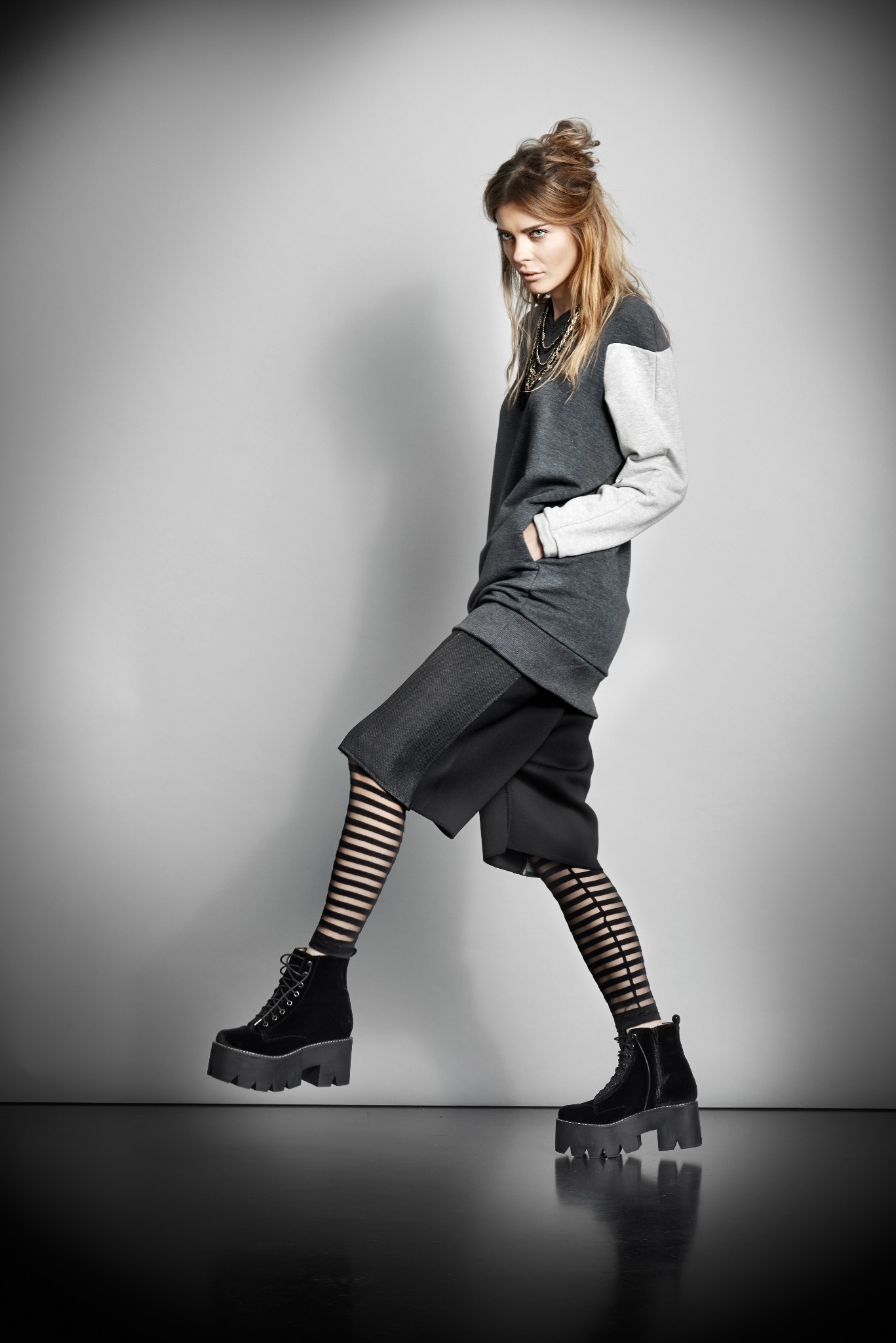 Modelka w szarej bluzie z kontrastowymi rękawami, krótkich spodenkach, rajstopach w paski i ciężkich butach na platformie, pozująca na szarym tle.